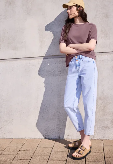 Eine Frau lehnt an einer grauen Betonwand und trägt ein Cap, braunes Shirt, hellblaue Jeans und Sandalen.